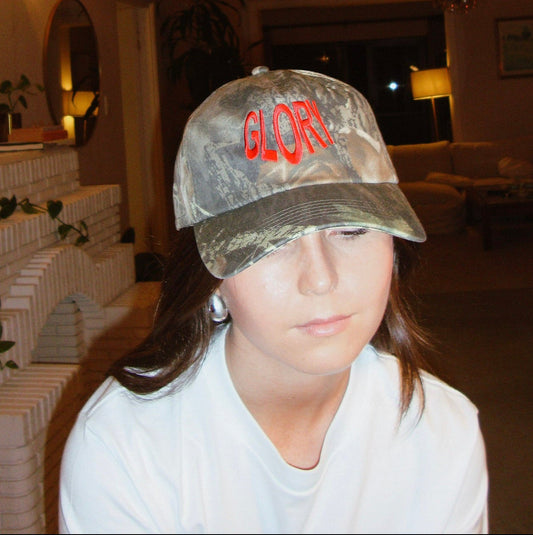 A person wearing a camouflage baseball cap with red embroidered letters 'GLORY' on the front, photographed indoors in a dimly lit living room.